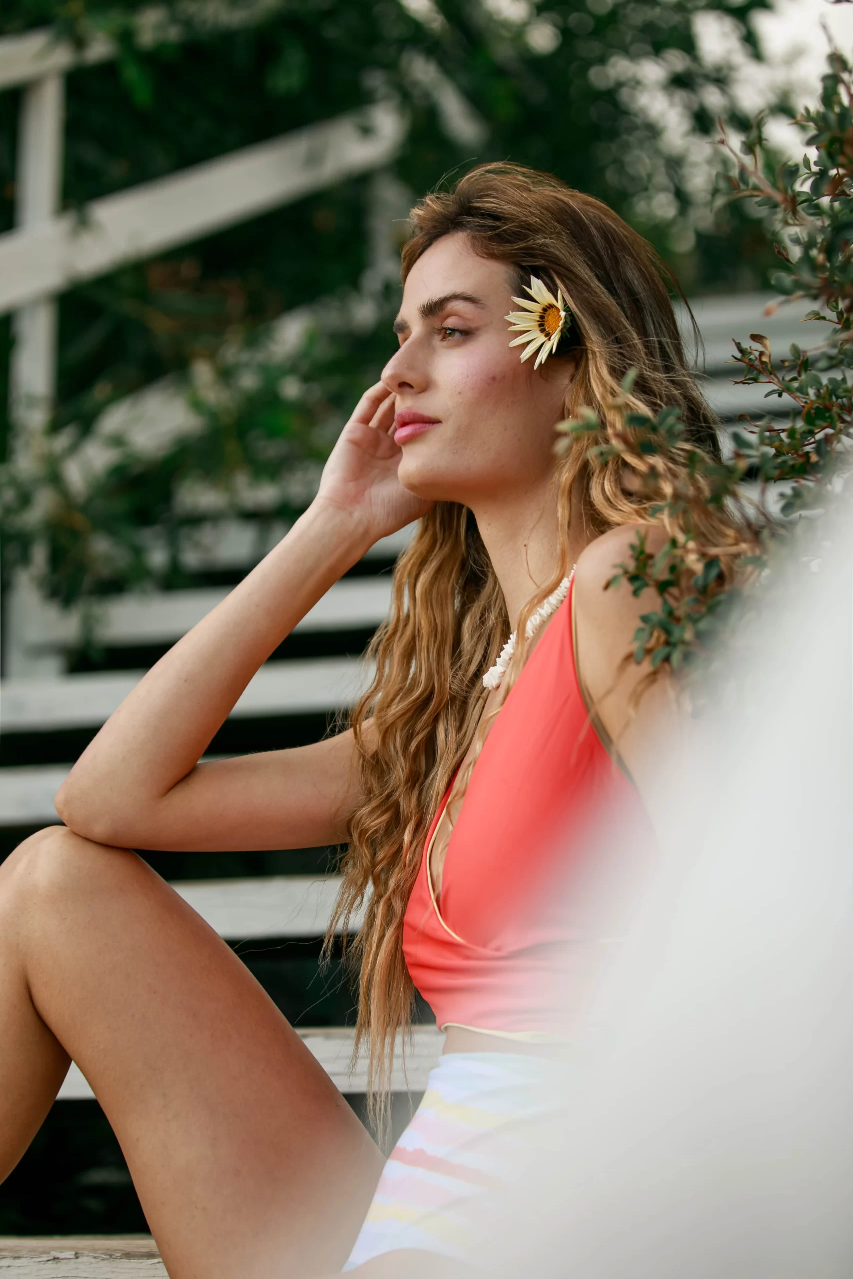 Hübsche junge Frau sitzt in einem Korallfarbenen Badeanzug auf einer Holztreppe. Sie trägt eine Blume in den Haaren. Man sieht sie im Profil.
